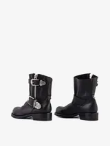Bota corta de cuero negro, estilo motorista, con caña baja y punta redonda. Presenta cierre lateral de cremallera y tiras ajustables con hebillas metálicas decorativas de estilo western.