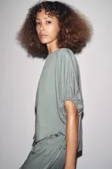 Blusa de cuello redondo color verde, confeccionada en hilatura con mezcla de lyocell. Manga corta abullonada ajustable con lazadas. Bajo redondeado.