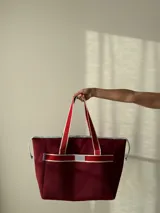 Bolso de mano color borgoña, confeccionado en neopreno, con cierre superior y asas de mano en cinta blanca y roja. Incluye monedero, bolsillo interno y externo con cierre, compartimentos internos y correa larga opcional.