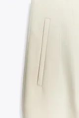 Pantalón de vestir color crema, de tiro alto, corte recto y largo cropped. Presenta bolsillos laterales de ojal y cierre frontal con cremallera, botón interior y gancho metálico.