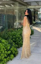 Vestido largo sin mangas, color dorado, con base beige y diseño de encaje o brocado con brillo. Corte ajustado al cuerpo que cae recto hasta el suelo.