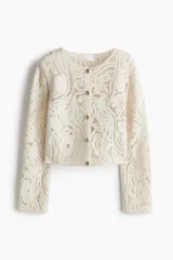 Saco corto color crudo, tejido al crochet con diseño floral. Tiene cuello redondo, mangas largas y cierre frontal con botones dorados.