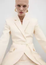 Blazer color crema con cuello de solapa, manga larga con puños abotonados, cinturón con hebilla y bolsillos delanteros con solapa.