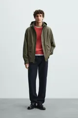 Parka verde militar de tejido técnico ligero resistente al viento, con cuello alto, capucha y manga larga con puños ajustables. Tiene bolsillos de plastrón con solapa en la cadera, bolsillo interior y bajo ajustable con elásticos. El cierre frontal es con cremallera oculta por solapa con botones a presión.