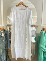 Vestido largo de lino blanco, de corte amplio, con cuello redondo y mangas cortas con botón. Presenta una hilera de botones a lo largo del frente.