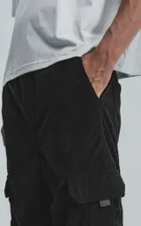 Pantalón cargo de pana negra, corte ancho y tiro alto con elástico en la cintura. Presenta bolsillos cargo amplios a los lados de las piernas, uno de ellos con un pequeño detalle de etiqueta o cierre.