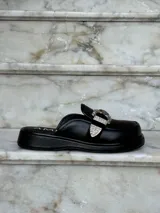 Zuecos negros de cuero con suela gruesa, detalle de tira de gamuza con hebilla metálica plateada y logo "AMÉN" estampado en la plantilla.