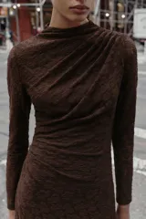 Vestido midi de manga larga, color marrón oscuro, confeccionado en tejido de encaje o punto con patrón floral sutil. Presenta cuello perkins y drapeado asimétrico en el torso con frunces laterales.