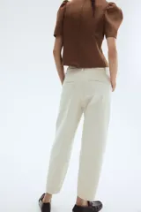 Jean tobillero de denim de algodón rígido color beige claro, con corte estándar de cadera a muslo y piernas con volumen de diseño curvo que se ciñen en el bajo. Presenta tiro alto, cierre con botón, bolsillos al bies, bolsillos decorativos traseros y pliegues delanteros y traseros.