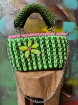Cartera tejida a crochet en hilo de algodón color verde con detalles en rosa y aplique de flor amarilla con lentejuelas. Tiene doble asa corta y correa larga de cadena metálica.