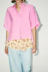Camisa rosa de cuello solapa y manga corta con vuelta. Bajo asimétrico con aberturas laterales. Cierre frontal con botones ocultos por solapa.