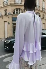 Camisa lila de cuello redondo y manga larga con volantes en el mismo tejido. Cierre en la espalda con aberturas y botones.