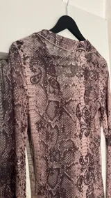 Vestido largo de gasa transparente con estampado de piel de serpiente en tonos marrones. Tiene cuello camisero, mangas largas acampanadas y abertura frontal con cordones.