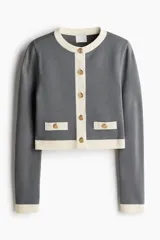 Cárdigan corto de punto fino color gris con cuello redondo y botones metálicos dorados. Tiene bolsillos insertados decorativos con botón adelante y ribete en color crema en cuello, cierre, bolsillos y puños.