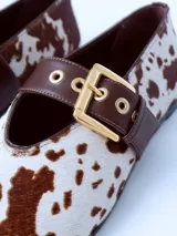 Zapatos bajos con estampado animal de vaca en blanco y marrón, con correa marrón y hebilla dorada.