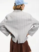 Blazer gris a rayas verticales blancas con lurex, con cuello de solapa, cierre con botón único y tajo en la espalda. Tiene un bolsillo de ojal en el pecho y bolsillos con solapa a la altura de la cadera.