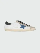 Championes urbanos Golden Goose modelo Super Star, color blanco con detalles en gris, negro y azul. Confeccionados en cuero y gamuza, con efecto desgastado y estrella azul en el lateral.