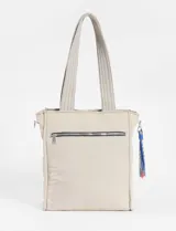 Bolso tipo shopper acolchado color beige claro, con costuras verticales onduladas en hilo azul. Posee dos asas de hombro y un pequeño accesorio colgante con cordones de colores en uno de los laterales. Presenta un logo cuadrado negro en el centro frontal.