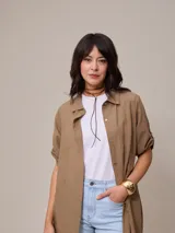 Vestido camisero largo color marrón claro, confeccionado en lino y algodón. Tiene cuello camisero, mangas 3/4 con botón para sujetar, cierre frontal con botones y corte recto. Se usa sobre una remera blanca y con un collar de cuero marrón.