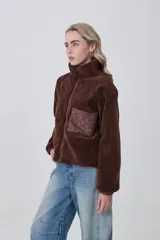 Campera de poliéster tipo sherpa en color natural, con diseño holgado, cuello alto y cierre frontal completo. Presenta un bolsillo de parche con cierre en el pecho y mangas largas con puños elásticos.