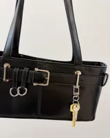 Cartera de hombro negra, de forma rectangular y acabado liso. Presenta detalles de hebillas plateadas y tiras decorativas en el frente, junto con un dije colgante en forma de llave dorada.