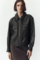 Cazadora negra de cuero con cuello solapa, manga larga con trabilla y bolsillos de parche con solapa en el delantero. Cierre frontal con cremallera y botones a presión ocultos por solapa.