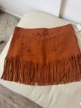 Pollera corta de gamuza color terracota/óxido, con detalle de costuras y apliques de cordones cruzados en la parte superior. El ruedo presenta flecos largos que aportan movimiento.