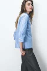 Camisa celeste a rayas verticales blancas, de cuello solapa y manga tres cuartos. Tiene aberturas laterales en el ruedo y cierre frontal con botones ocultos.
