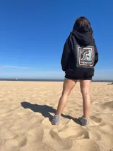 Canguro negro con capucha y cordones ajustables. Estampa en la espalda con el texto "Chema Surf Trip" y dibujos de un sol, una tabla de surf, una carpa y otros elementos relacionados con el surf.