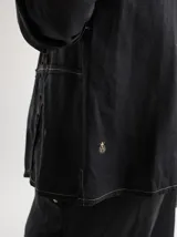 Camisa negra de lino con corte femenino, cuello clásico, cierre frontal con botones y mangas 3/4 con puños. Presenta detalles de frunces en el frente.