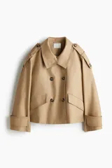 Gabardina corta color beige con doble botonadura, cuello clásico, charreteras en los hombros, bolsillos delanteros con solapa, mangas anchas con trabillas ajustables y pliegue en la espalda.