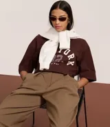 Remera de algodón color marrón oscuro con cuello redondo y mangas cortas, de corte oversized. Presenta un estampado frontal con el texto "NEW YORK 82" en tipografía universitaria y la frase "STATE OF MIND" en la parte inferior.