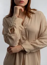 Vestido camisero midi color beige, de corte holgado, con cuello camisero, mangas largas con puños abotonados y aberturas laterales en el ruedo.