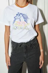 Remera blanca de algodón con cuello redondo y mangas cortas. Estampado frontal con un caballo azul, rosas rosadas y texto bordado en hilo amarillo.
