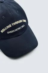 Gorra azul marino con visera y bordado frontal con la frase "Going Through Time Garden District, NOLA" en color crema. Ajuste trasero con hebilla metálica.