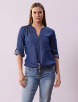 Blusa de tencel azul con cuello a la base y escote en V, abertura central con botones, bolsillos delanteros, mangas 3/4 regulables con presilla y botón, y terminación de ruedo con nudo.