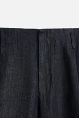 Pantalón de denim azul oscuro, corte wide fit, largo cropped, con pliegues frontales, bolsillos laterales y traseros asimétricos. Cierre frontal con cremallera y gancho metálico oculto.