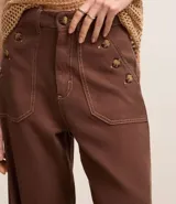 Pantalón wide leg color marrón chocolate, confeccionado en sarga de algodón y lyocell, con cintura alta, trabillas, cierre con botón y cremallera, y bolsillos tipo carpintero. Presenta botones color carey en los bolsillos delanteros.