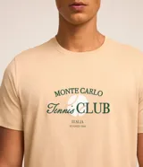 Remera color arena de algodón, con cuello redondo y mangas cortas. Estampa frontal con el texto "Monte Carlo Tennis Club" en color verde.