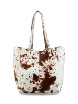 Bolso tote de cuero con pelo, estampado de vaca en blanco y marrón. Tiene asas de cuero marrón y un pequeño parche de cuero marrón con logo grabado en el frente.