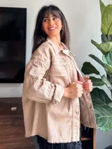 Chaqueta color beige con bordado calado, cuello camisero, cierre frontal con botones, mangas largas con puños abotonados, bolsillos de parche en el pecho y ruedo desflecado.