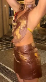 Top halter de cuero marrón con diseño de chaleco vaquero, ajustado al cuello y con cierre frontal mediante hebillas doradas. Presenta bordados en hilo dorado con motivos de llamas o plumas estilizadas en el frente.