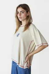 Remera beige de corte holgado con cuello redondo y mangas cortas. Presenta un pequeño estampado de un sol azul en el centro del pecho y costuras en color rojo en cuello, mangas y ruedo.