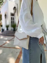 Cartera tipo sobre color crudo, confeccionada en cuero, con correa larga de cuero marrón y costuras decorativas en contraste.