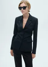 Americana de estilo traje color negro, con cuello pico, cierre delantero con botones, y dos bolsillos de ribete.