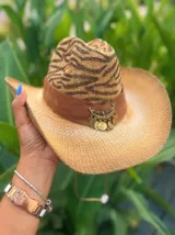 Sombrero de ala ancha tejido en hilo de coco y sintético, color marrón claro. La copa está cubierta con encaje naranja quemado con patrón floral. Tiene una cinta de cuero marrón oscuro y un aplique decorativo de hoja de gamuza con incrustaciones de pedrería.