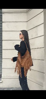 Bolso tipo hobo color tostado, hecho de cuero con flecos largos en el frente y dos bolsillos con cierre. Tiene una manija regulable con hebillas metálicas.