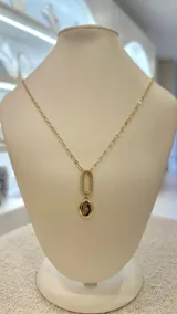 Collar largo dorado con cadena de eslabones y dije con forma de óvalo con textura trenzada, del que cuelga un dije redondo con una circonia engarzada.