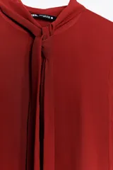 Blusa de cuello subido color ladrillo, confeccionada en hilatura de viscosa 100%. Manga larga con pliegues y abullonada. Detalle aplicación de lazo en cuello ajustable con lazada. Cierre frontal con botones.