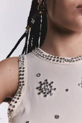 Musculosa color crudo de punto canalé, con cuello redondo y sisas ribeteadas con strass. Presenta apliques de strass con forma de flor y tachas plateadas dispersas por toda la prenda.